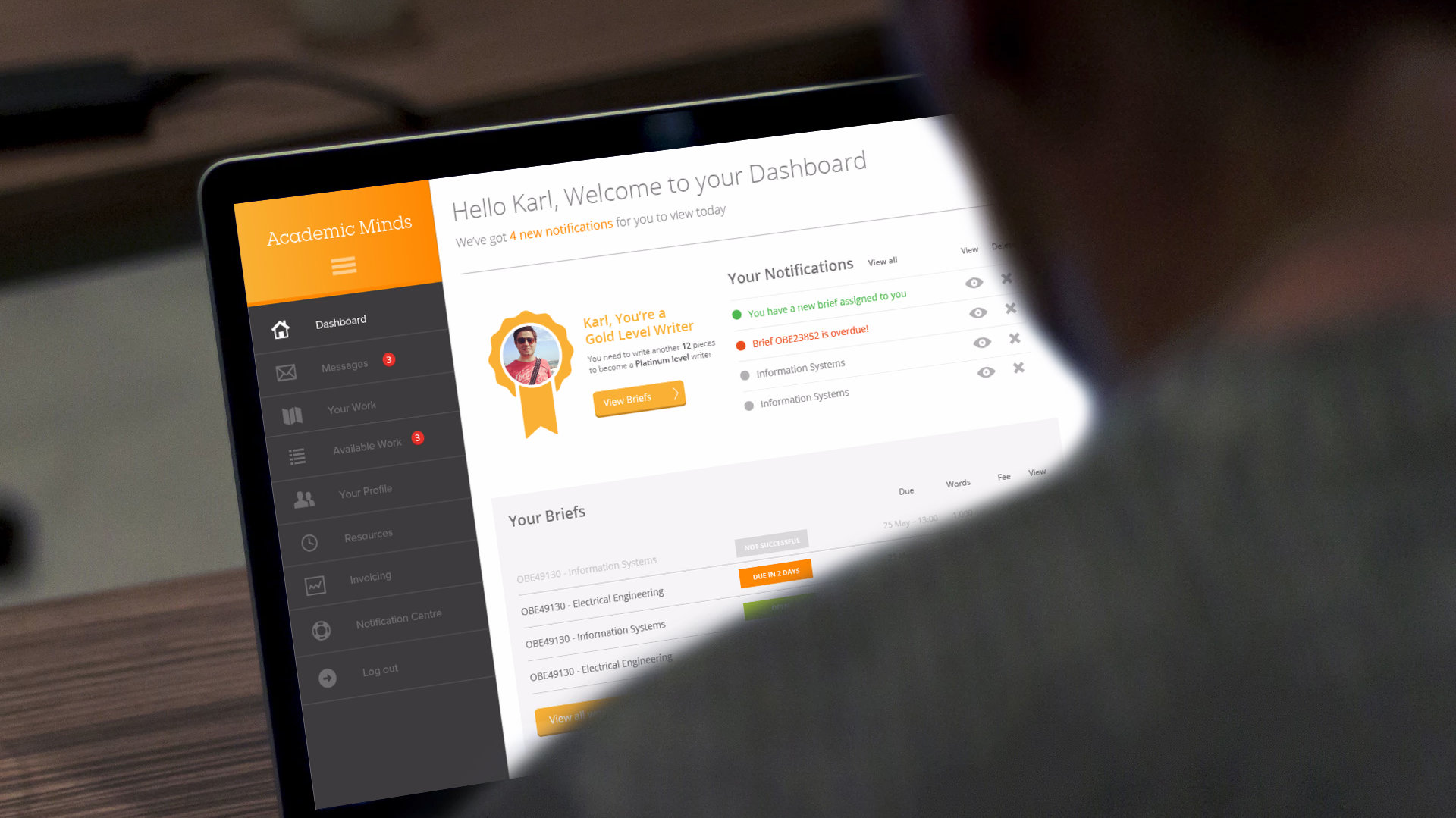 The portal dashboard on a laptop, seen over the writer's shoulder. A dark sidebar lists Dashboard, Messages, Your Work, Available Work, Your Profile, Resources, Invoicing and Notification Centre. The main area says 'Hello Karl, Welcome to your Dashboard', shows the writer's Gold Level badge, a list of notifications and a table of briefs with their status.